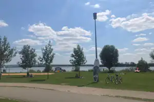 Grüne Campingwiese mit Fahnenmast, Fahrrädern und mehreren Sitzgelegenheiten, im Hintergrund ein Gebäude und Bäume.