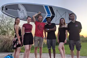Gruppe von Campern vor einem großen Surfboard am Campingplatz, im Hintergrund Wiese und Bäume.
