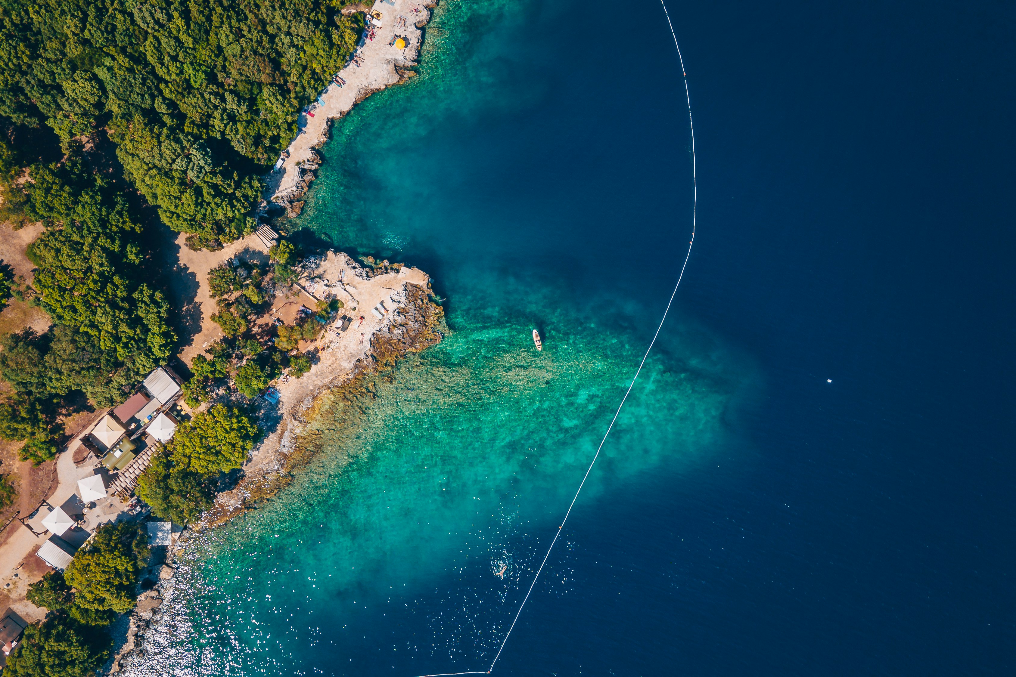 Luftaufnahme einer türkisfarbenen Bucht mit Strandabschnitten, Booten und einem kleinen Campingbereich am Ufer.