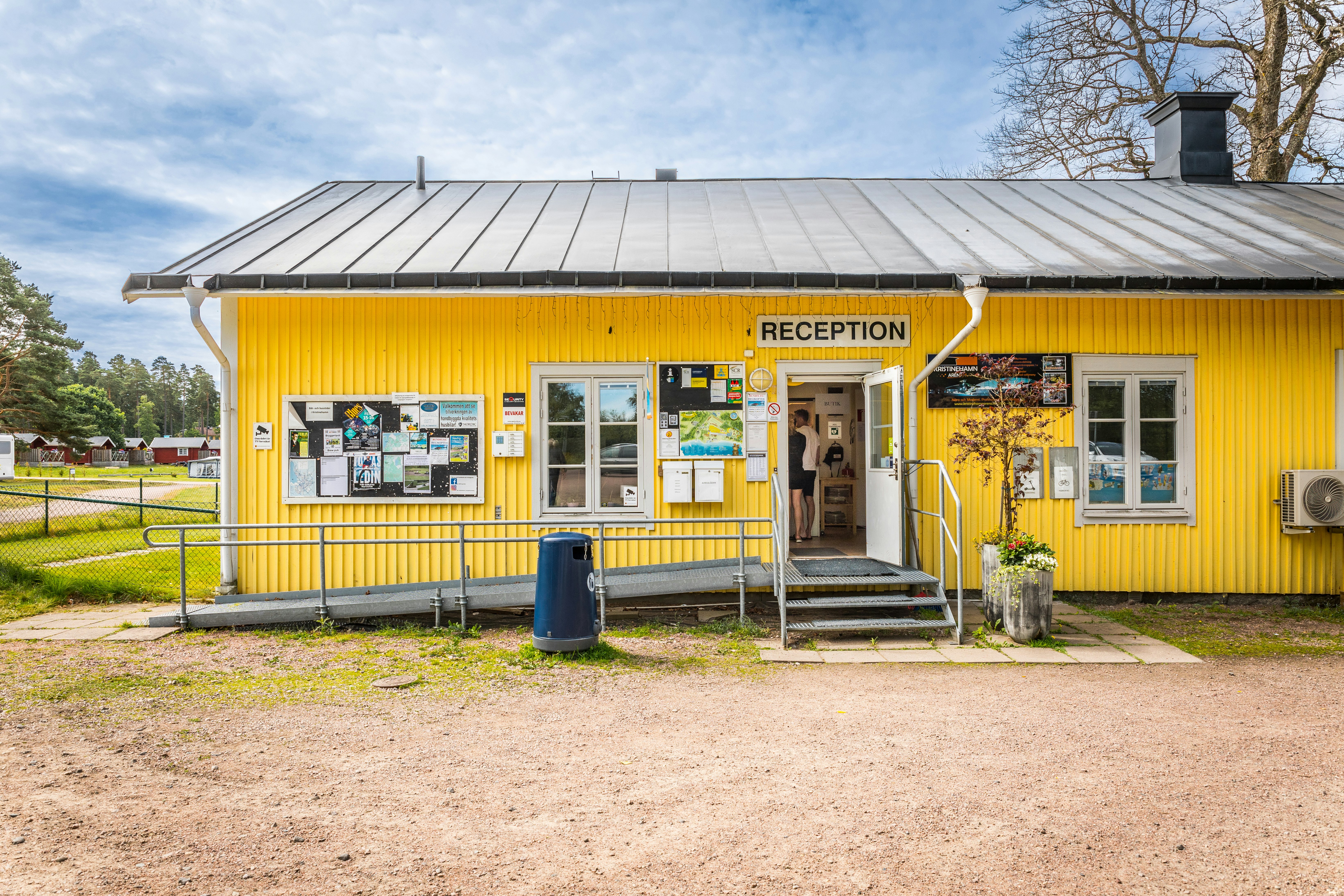 Gelbes Gebäude mit Rezeption am Campingplatz, Eingang mit Stufen, Aushangtafeln und Briefkasten, davor Kiesweg und Rasen.