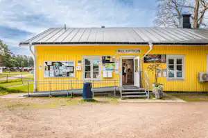 Gelbes Gebäude mit Rezeption am Campingplatz, Eingang mit Stufen, Aushangtafeln und Briefkasten, davor Kiesweg und Rasen.