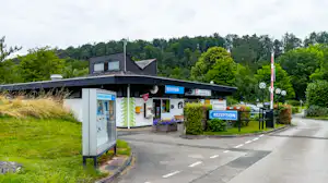 Rezeption eines Campingplatzes mit Schildern, Eingangstür, Sitzbank und Weg vor dem Gebäude, umgeben von Bäumen und Rasen.