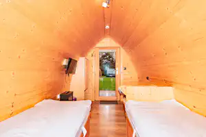 Innenansicht einer Holzhütte mit zwei Betten, schmalem Gang und Blick durch die Tür auf den Garten.