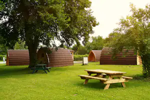 Holzhütten auf grünem Rasen, dazwischen ein Picknicktisch und Bäume im Hintergrund.