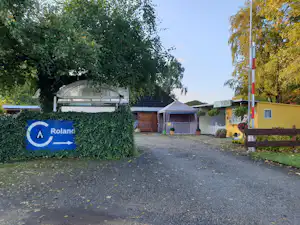 Campingplatz-Eingang mit Schild „Roland“, Kiesweg, Hecken und gelbem Gebäude im Hintergrund.