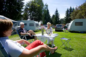 Campingplatz mit Wohnwagen und Wohnmobilen, Rasen und Tisch mit Blumenstrauß, mehrere Personen sitzen im Gartenstuhl.