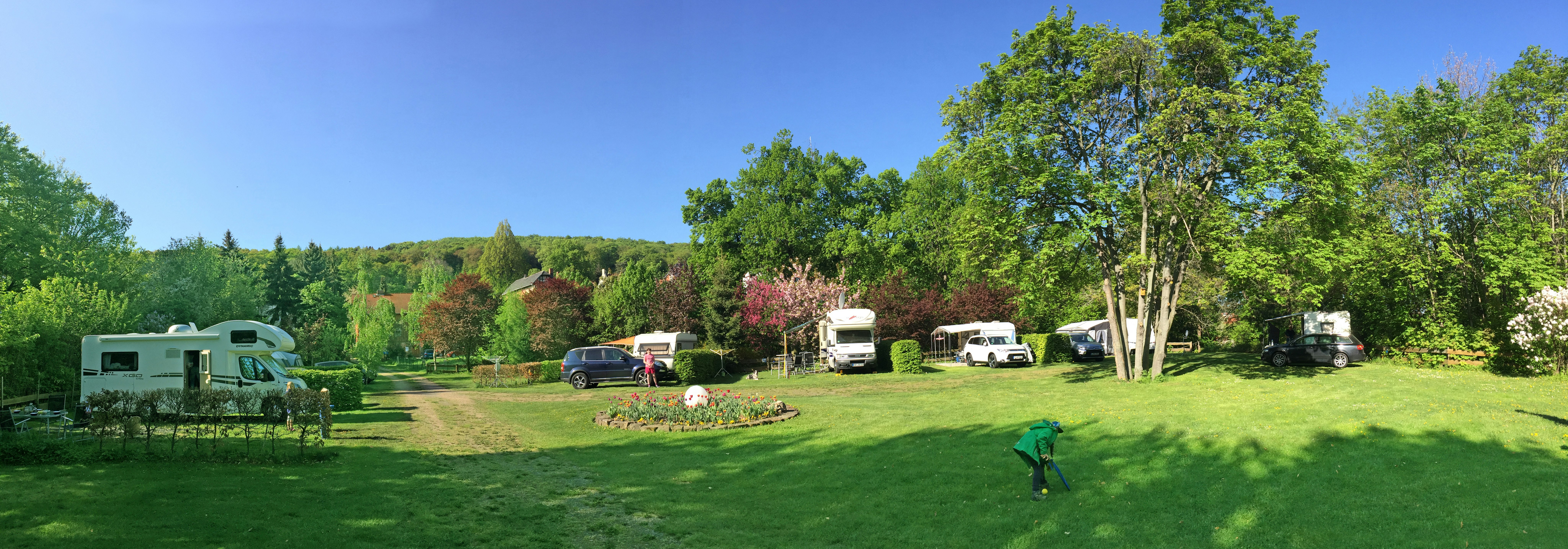 Campingwiese mit Wohnmobilen und Wohnwagen, Bäumen und Hecken im Hintergrund, grüner Rasen und freier Blick ins Tal.