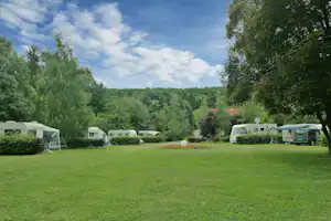 Grüne Campingwiese mit mehreren Wohnwagen, einem Wohnmobil und Bäumen am Rand, im Hintergrund dichter Wald.