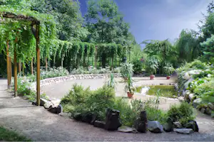 Campinggarten mit Kiesweg, Beeten und Terrassenstufen, umgeben von Bäumen und Sträuchern.