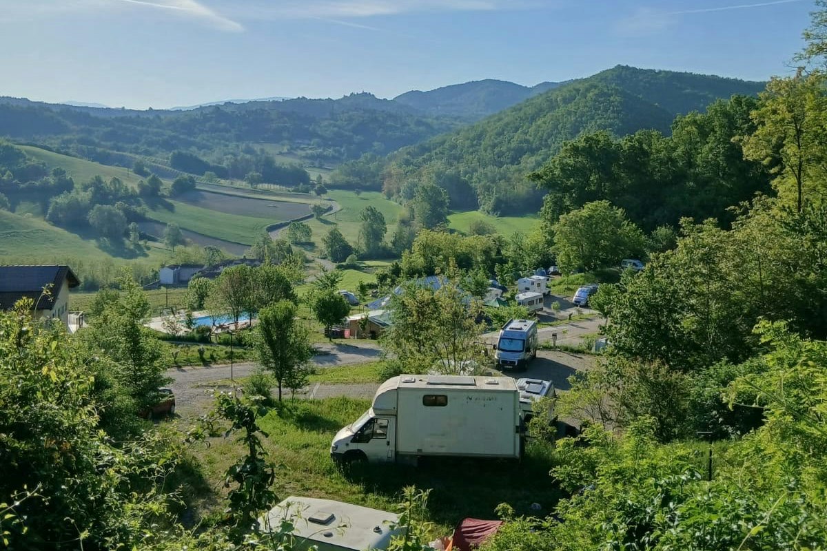 Wohnwagen und Campingplatz im Tal, umgeben von grünen Bäumen, Hügeln und einem Flusslauf unter blauem Himmel.