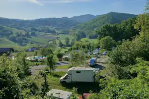 Wohnwagen und Campingplatz im Tal, umgeben von grünen Bäumen, Hügeln und einem Flusslauf unter blauem Himmel.