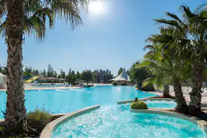 Campingpool mit türkisfarbenem Wasser, Palmen und weißen Liegen am Beckenrand, im Hintergrund ein weißes Zelt.