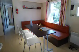 Wohnbereich mit brauner Eckcouch, Esstisch und Stühlen, Fenster mit Vorhängen und Blick ins Grüne, Feuerlöscher an der Wand.