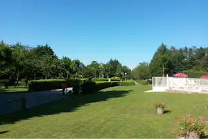 Grüner Rasen und Hecken am Campingplatz, gepflasterter Weg, weiße Zäune und rote Sonnenschirme neben einem Sitzbereich.