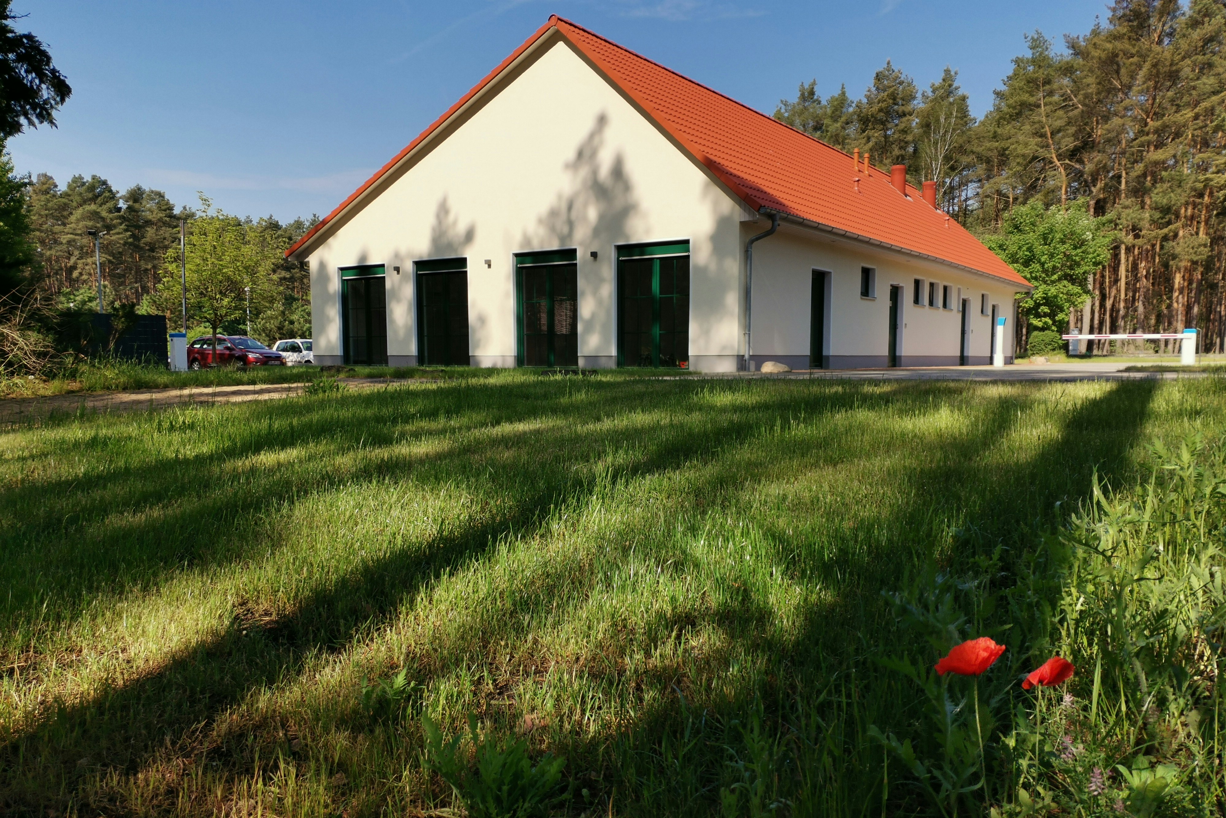 Weißes Campinggebäude mit rotem Dach, grüner Rasenfläche davor und Bäumen im Hintergrund, seitlich Fenster und Eingang.