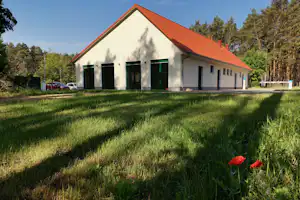 Weißes Campinggebäude mit rotem Dach, grüner Rasenfläche davor und Bäumen im Hintergrund, seitlich Fenster und Eingang.