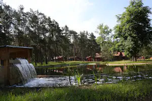 Teich mit kleinem Wasserfall, umgeben von Gras und Schilf, dahinter Bäume und Holzhäuser auf dem Campingplatz.