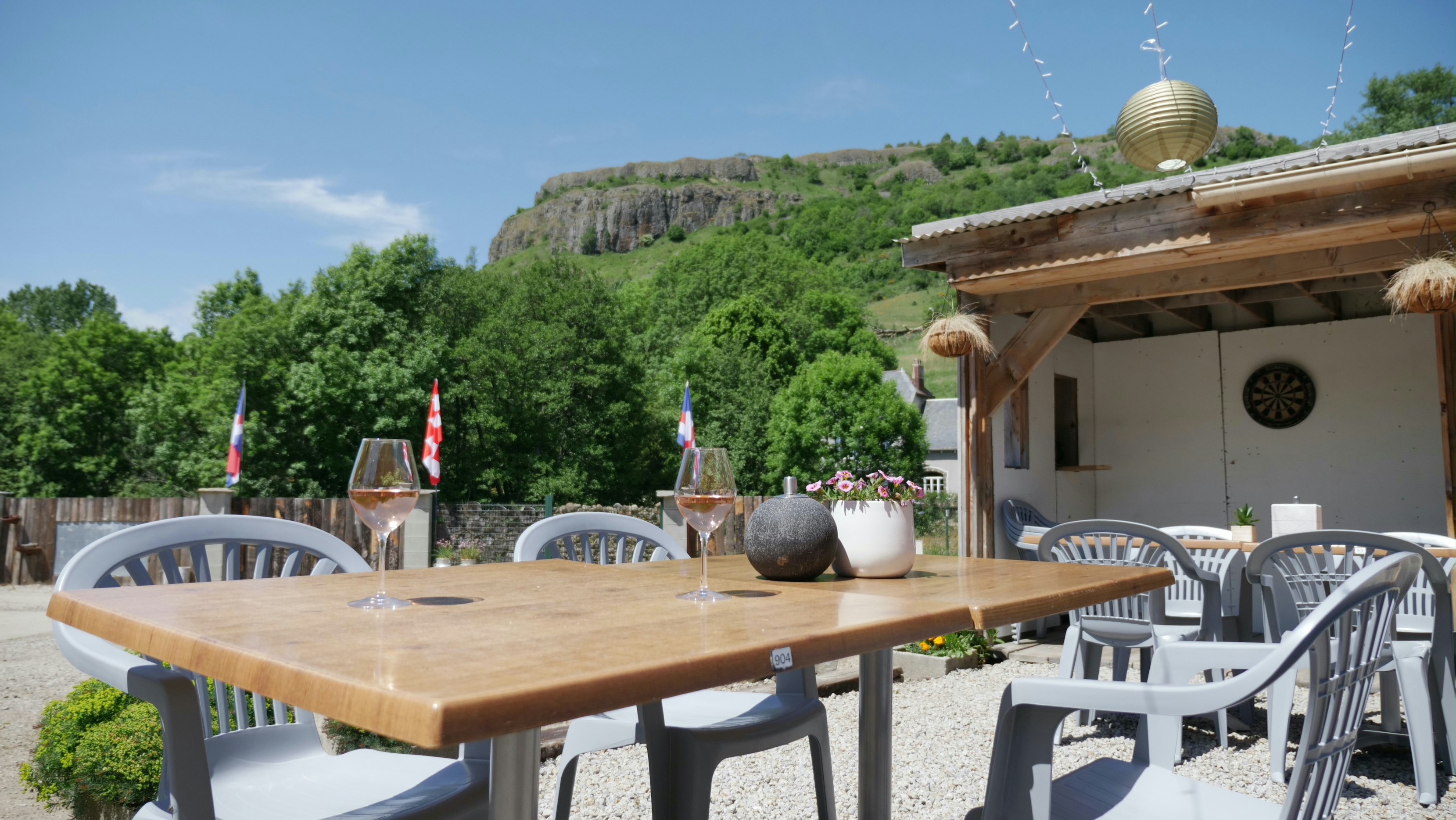 Holztisch mit Stühlen auf einer Terrasse, daneben Deko und Laternen, im Hintergrund Bäume und eine Felswand.