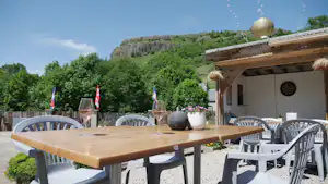Holztisch mit Stühlen auf einer Terrasse, daneben Deko und Laternen, im Hintergrund Bäume und eine Felswand.