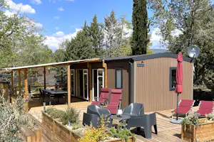 Bungalow mit Holzterrasse, Sitzgruppe und Sonnenschirm, umgeben von Büschen und Bäumen im Campingbereich.