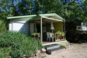 Mobilheim mit Veranda und Markise, davor weiße Gartenstühle und Holztisch, umgeben von Büschen und Kiesweg.