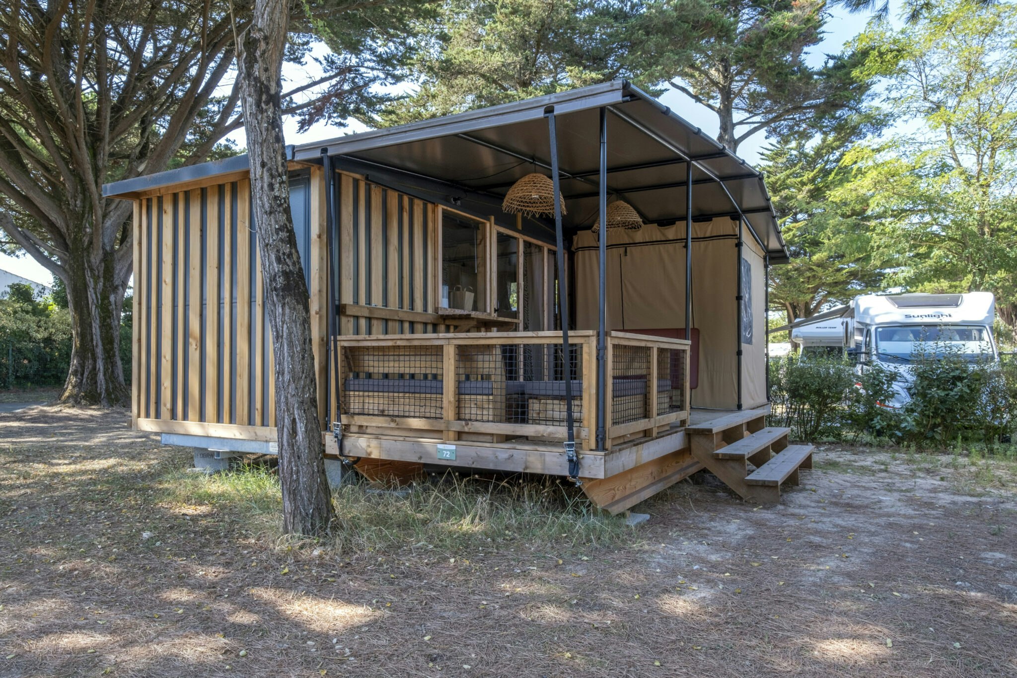 Holzunterkunft mit Veranda und Zeltplane, davor Stufen und Holzgeländer, im Hintergrund Bäume und ein Wohnmobil.