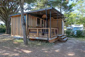 Holzunterkunft mit Veranda und Zeltplane, davor Stufen und Holzgeländer, im Hintergrund Bäume und ein Wohnmobil.