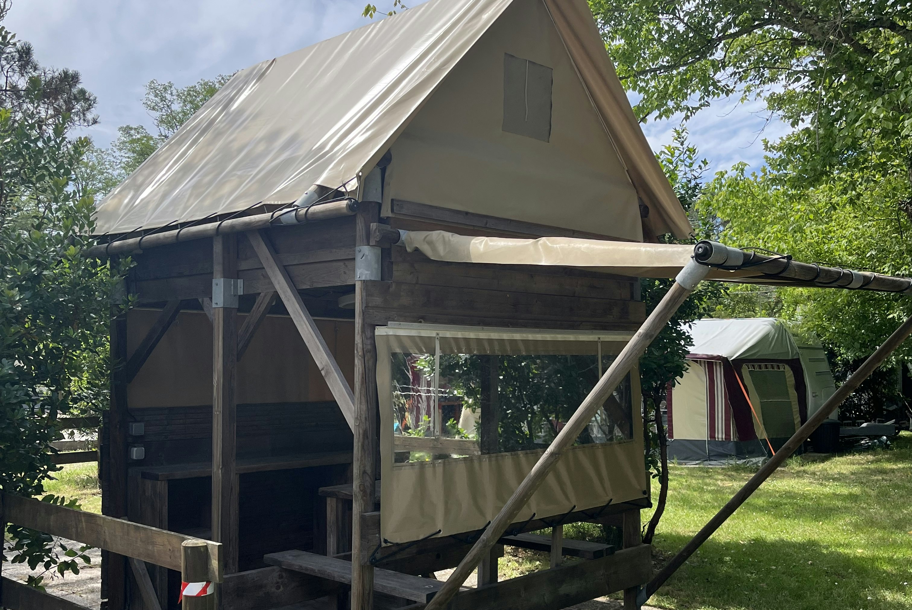 Zelt auf Stelzen mit Holzplattform, Vorhang und Leiter, im Grünen zwischen Bäumen auf einem Campingplatz.