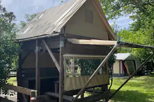 Zelt auf Stelzen mit Holzplattform, Vorhang und Leiter, im Grünen zwischen Bäumen auf einem Campingplatz.
