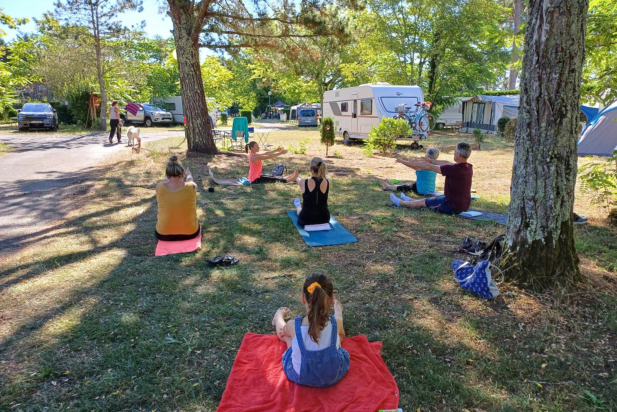 Campingplatz mit Wohnwagen und mehreren Personen beim Yoga auf Matten im Gras zwischen Bäumen.