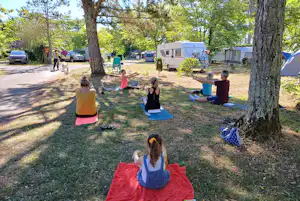 Campingplatz mit Wohnwagen und mehreren Personen beim Yoga auf Matten im Gras zwischen Bäumen.