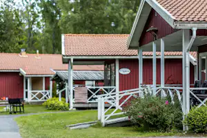 Campinghaus mit roter Holzfassade, überdachter Terrasse und weißen Geländern, im Hintergrund Bäume und Rasen.
