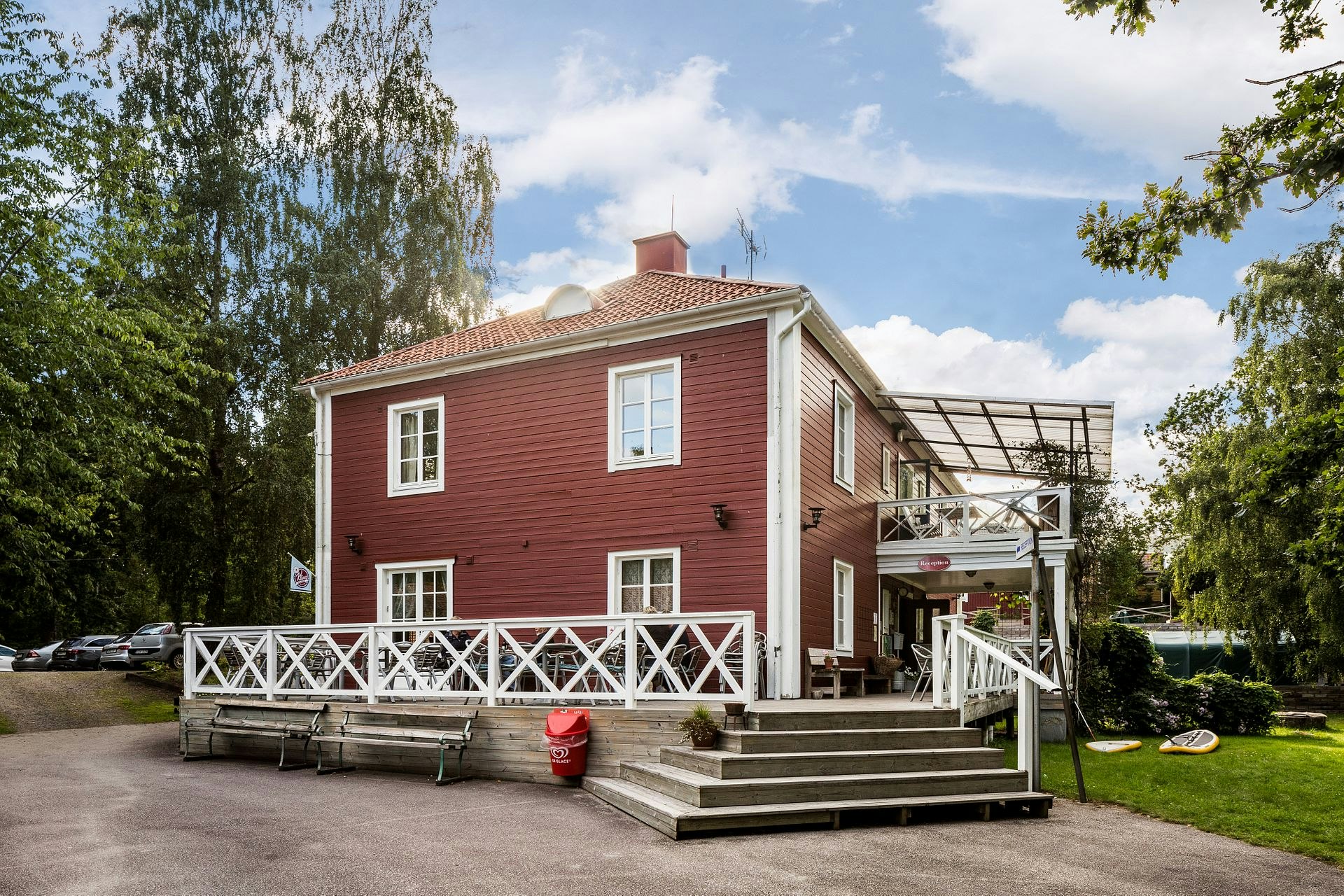 Rezeptionsgebäude mit roter Holzfassade, weißem Geländer und überdachtem Eingang, umgeben von Bäumen und Parkplatz.