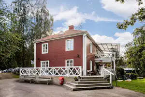 Rezeptionsgebäude mit roter Holzfassade, weißem Geländer und überdachtem Eingang, umgeben von Bäumen und Parkplatz.