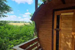 Holzhütte mit Tür und Terrasse am Campingplatz, umgeben von Büschen und Wiese, im Hintergrund Bäume und Feld.