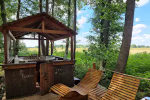 Holzhütte mit überdachter Terrasse, Grill und Sitzbänken auf einem Campingplatz im Wald mit Blick ins Grüne.