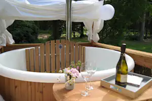 Privater Whirlpool auf einer Holzterrasse mit Flasche Wein, Gläsern und Blumenstrauß, umgeben von Zäunen und Bäumen.