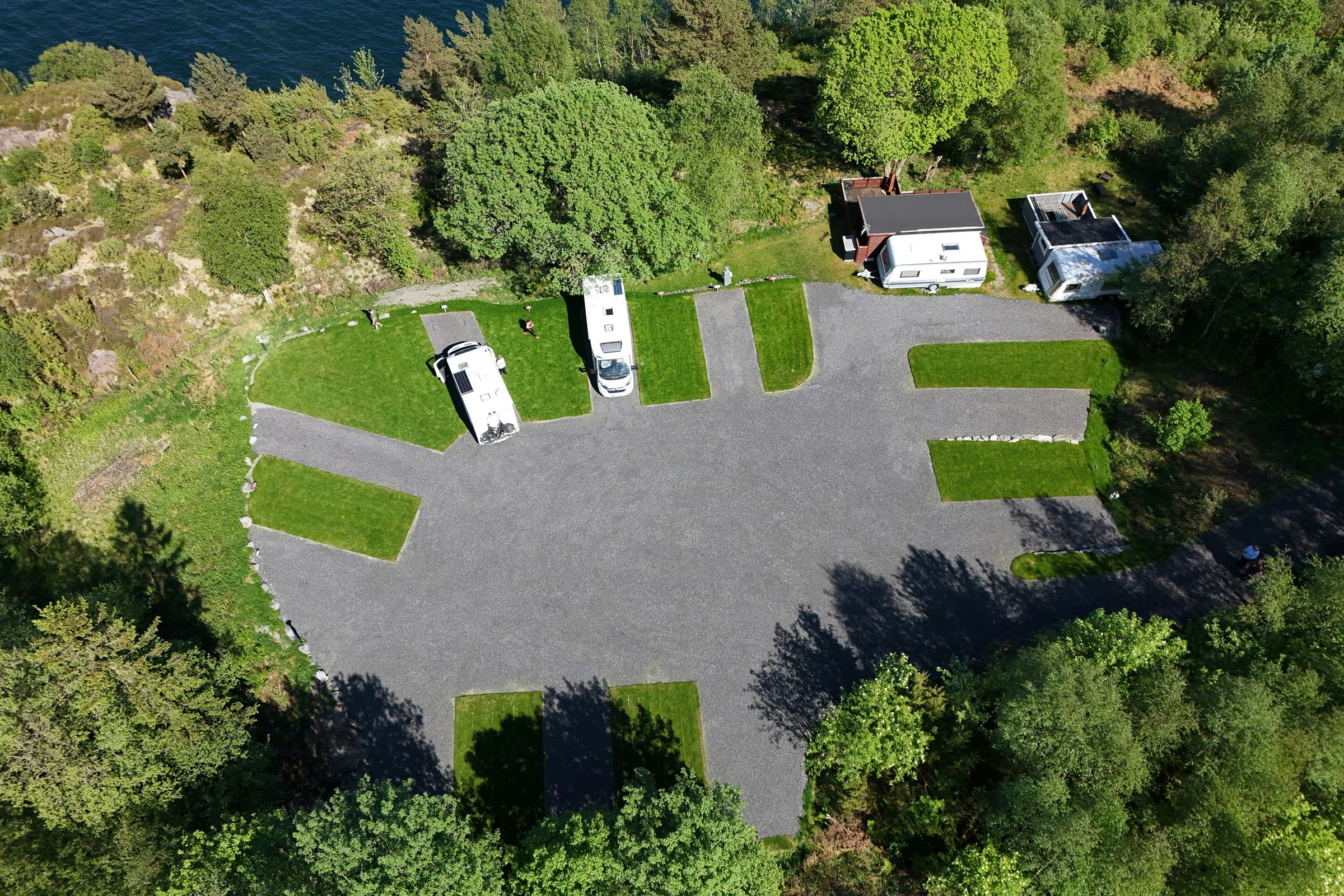 Luftaufnahme eines Campingplatzes mit Stellplätzen, Wohnmobilen und einem großen Asphaltbereich, umgeben von Bäumen und Grünflächen.
