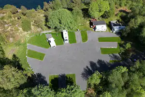 Luftaufnahme eines Campingplatzes mit Stellplätzen, Wohnmobilen und einem großen Asphaltbereich, umgeben von Bäumen und Grünflächen.