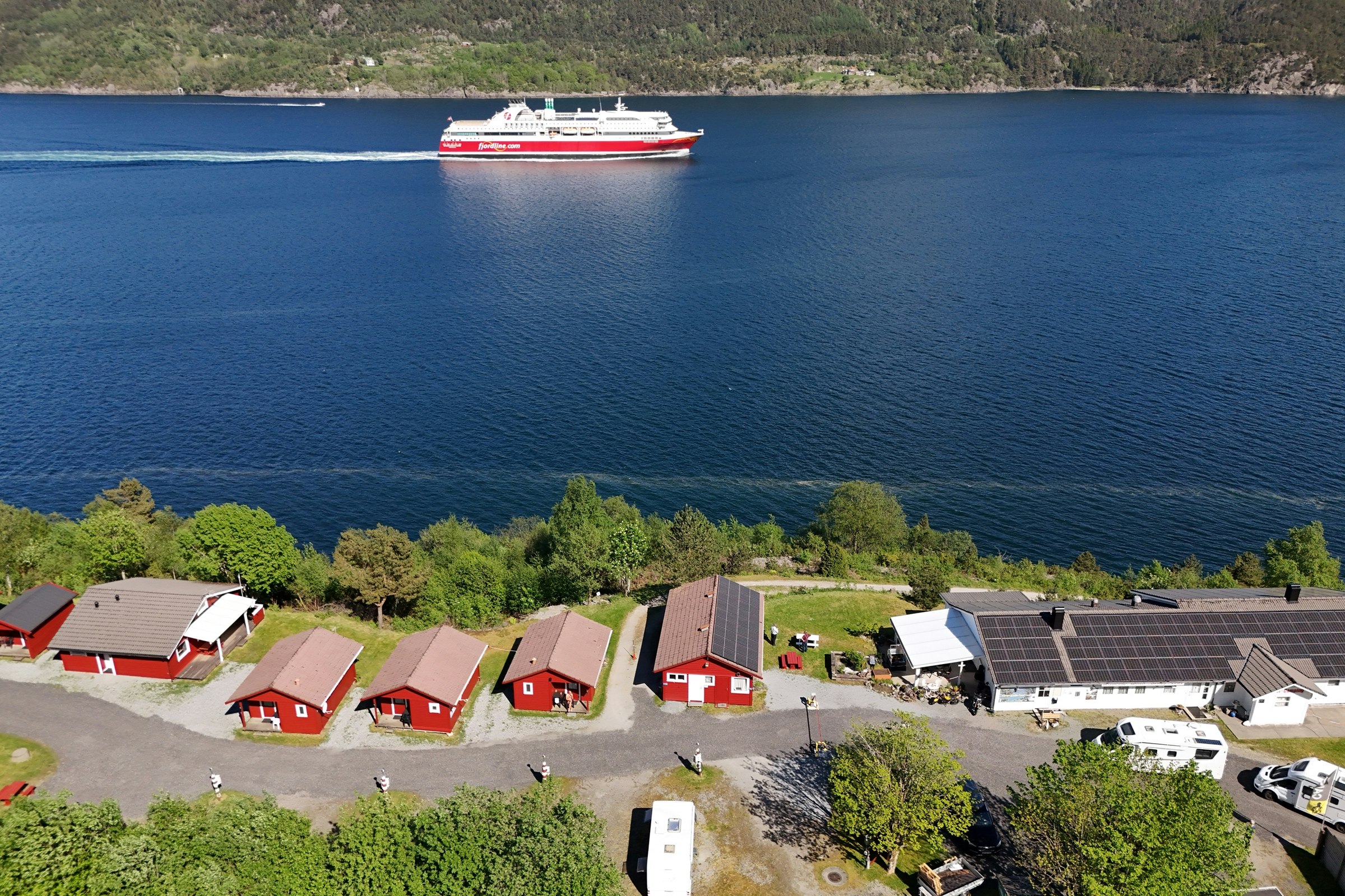 Blick auf ein Campinggelände am See mit roten Ferienhäusern, Stellplätzen und einem Schiff auf dem Wasser.