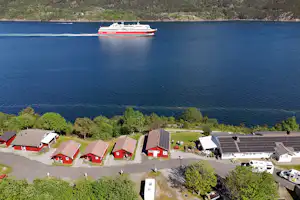 Blick auf ein Campinggelände am See mit roten Ferienhäusern, Stellplätzen und einem Schiff auf dem Wasser.