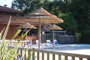 Camping-Terrasse mit weißen Liegestühlen und mehreren Strohdach-Schirmen vor Holzgebäuden, umgeben von Büschen und Bäumen.