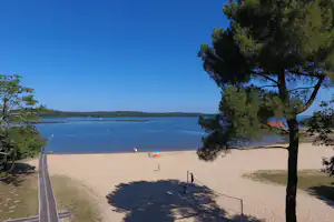 Sandstrand am See mit großen Bäumen im Vordergrund, Weg am Ufer und Blick über das Wasser.