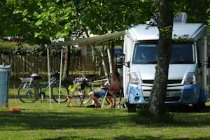Wohnmobil am Campingplatz mit Fahrrad, Sonnenschirm und Tisch unter Bäumen, daneben ein weiterer Stellplatz mit Wäscheleine.