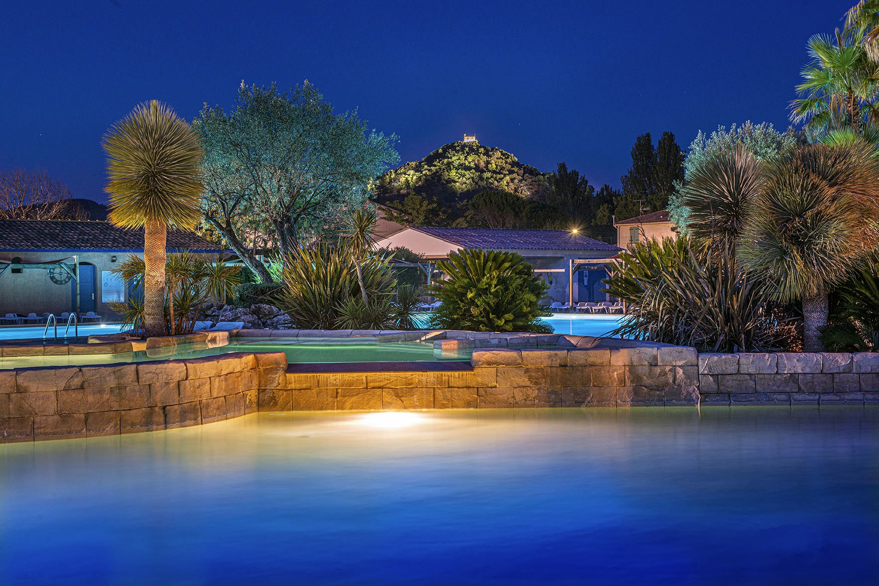 Beleuchteter Pool im Campingbereich bei Nacht, umgeben von Palmen, Steinmauer und Liegen am Wasserbecken.