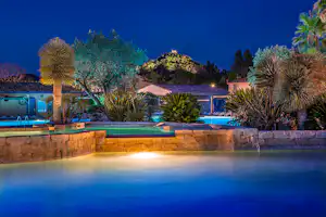 Beleuchteter Pool im Campingbereich bei Nacht, umgeben von Palmen, Steinmauer und Liegen am Wasserbecken.