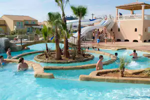 Schwimmbecken auf einem Campingplatz mit Palmen, Wasserrutsche und mehreren Personen im Wasser vor weißen Gebäuden.