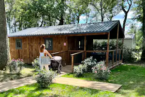 Holz-Cottage mit Veranda, großen Fenstern und Holzgeländer im Grünen, daneben Blumenbeete und ein Rollwagen.