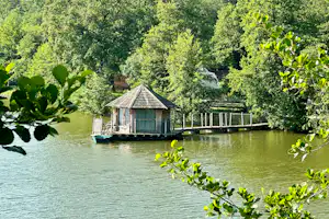Bootshaus auf einem Steg am See, umgeben von Bäumen und grünem Wasser, im Hintergrund weitere Gebäude.