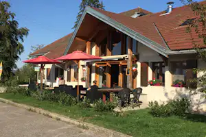 Campinghaus mit überdachter Terrasse, roten Sonnenschirmen, Holzbalken und Sitzgruppe vor dem Eingang, umgeben von Grünpflanzen.